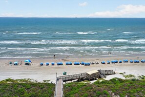 A beach with a wooden boardwalk leading to a building, and several blue umbrellas set up along the shore. - Sandpiper 1005: Oceanfront 3BR condo with pool, hot tub, and beach access in Port Aransas. (Port Aransas)