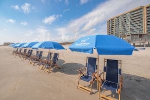 A beach with rows of blue beach chairs and umbrellas, a high-rise building in the background. - Sandpiper 1005: Oceanfront 3BR condo with pool, hot tub, and beach access in Port Aransas. (Port Aransas)