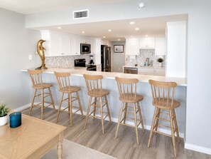 A modern kitchen with white cabinets, a central island with bar stools, and a dining table with chairs. - Sandpiper 1005: Oceanfront 3BR condo with pool, hot tub, and beach access in Port Aransas. (Port Aransas)