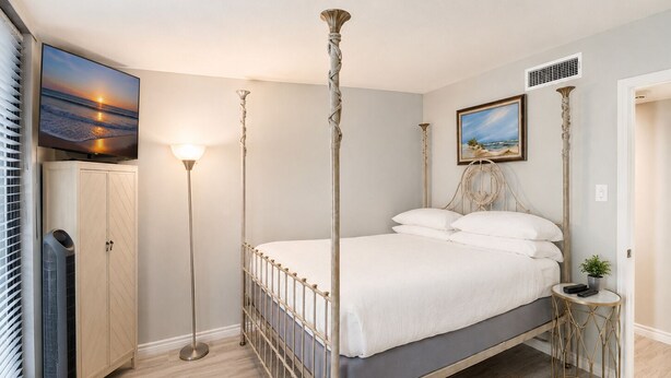 A bedroom with a bed, a television, a wardrobe, a floor lamp, and a painting on the wall. - Sandpiper 1005: Oceanfront 3BR condo with pool, hot tub, and beach access in Port Aransas. (Port Aransas)