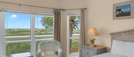 A hotel room with a bed, a chair, a table, and a lamp. There are large windows with curtains and a view of the beach.