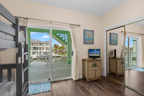 A room with a sliding glass door leading to a balcony, a television on a wooden cabinet, and two framed artworks on the wall. - Dolphins and Dunes | Private Boat Dock | Fishing Poles & Beach Gear | My Beach Getaways (Pensacola Beach)