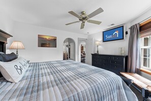 Ein Schlafzimmer mit Bett, Deckenventilator, Fernseher, Kommode, Fenster mit Vorhängen und einem Bild an der Wand.