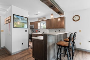 A modern kitchen with a central island, wooden bar stools, and a large clock on the wall. - Casa de Cozy - Resort Condo Minutes from Branson & Lake Taneycomo (Branson)