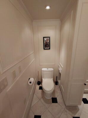 Een kleine, smalle badkamer met toilet, een ingelijste foto aan de muur en een rol toiletpapier op de vloer.
