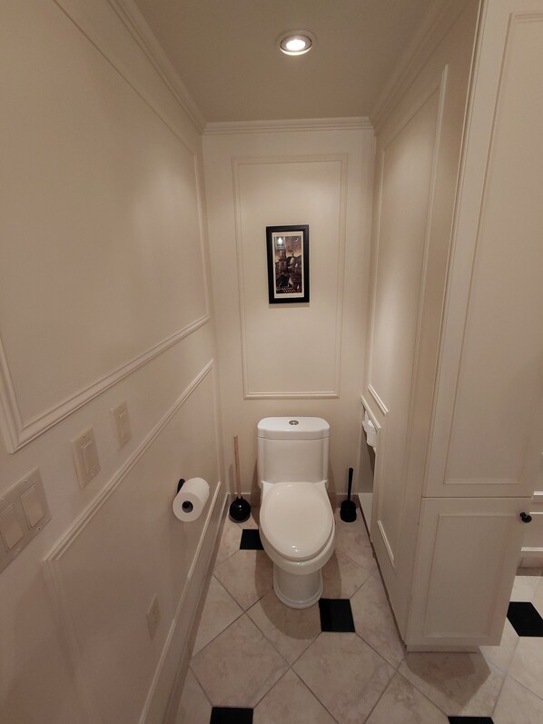 A small, narrow bathroom with a toilet, a framed picture on the wall, and a roll of toilet paper on the floor.