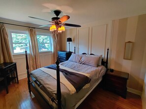 A bedroom with a four-poster bed, a ceiling fan, a nightstand, a dresser, and a window with curtains.