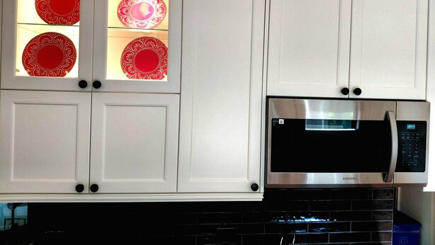 Una cocina con armarios blancos, un microondas de acero inoxidable y platos decorativos rojos en el interior de los armarios.