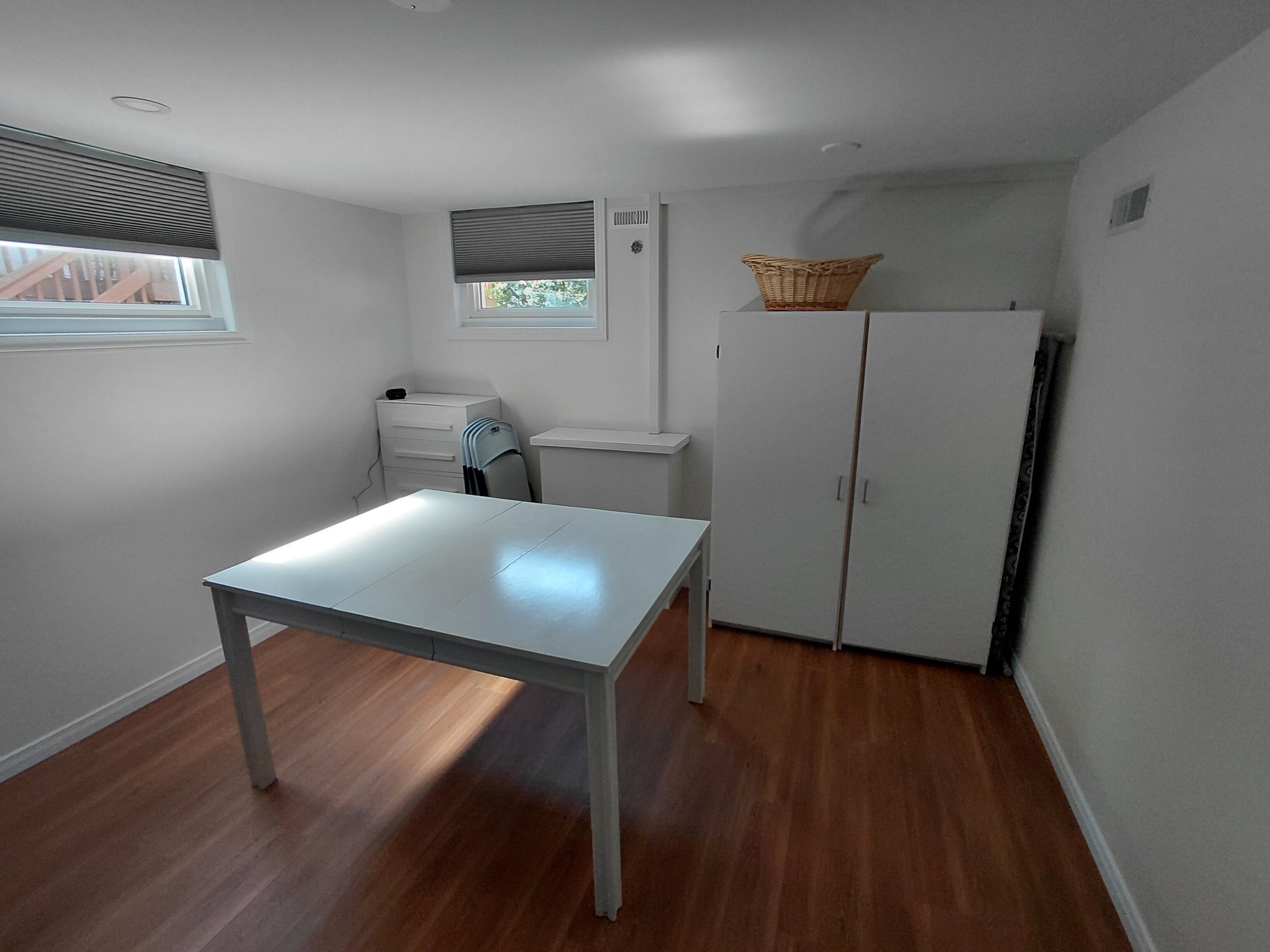 Une pièce avec une table blanche, une armoire blanche, une fenêtre avec des stores et un petit panier posé sur le dessus de l’armoire.