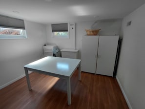 A room with a white table, a white wardrobe, a window with blinds, and a small basket on top of the wardrobe.