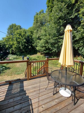 A wooden deck with a glass table and chairs, an umbrella, and a railing overlooking a garden.