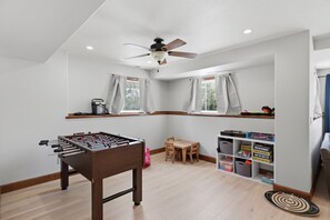 Ein Zimmer mit Kicker, Deckenventilator, einem fenster mit Vorhängen und einem Regal mit Büchern und Spielzeug.