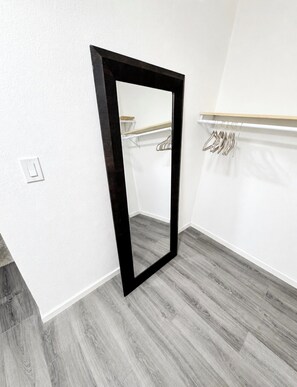 Um quarto com um espelho emoldurado a preto, uma prateleira de madeira com cabides e um interruptor de luz na parede.