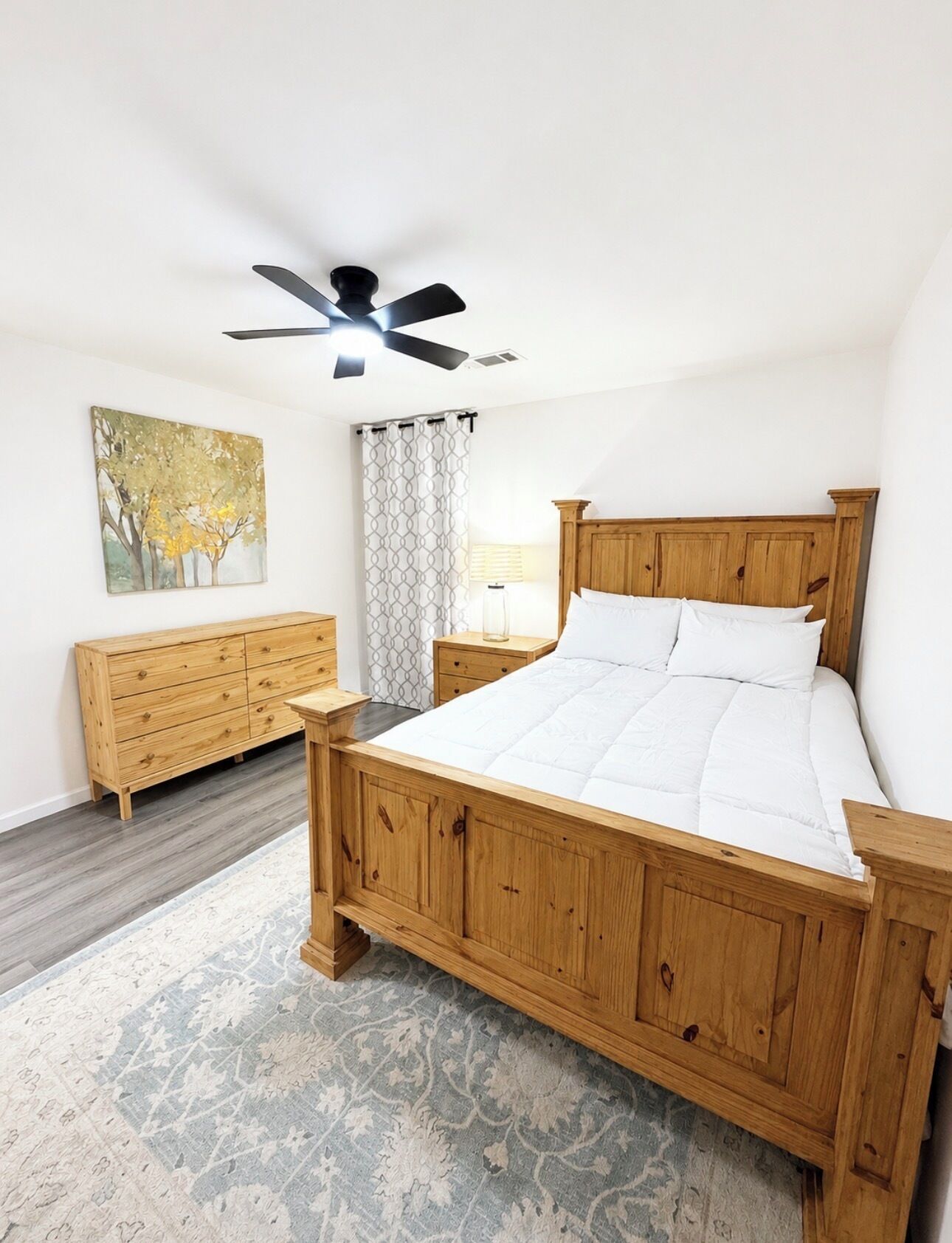 Ein Schlafzimmer mit einem hölzernen Bett, einem Deckenventilator, einem Nachttisch, einer Kommode, einem gemusterten Teppich und einem Bild an der Wand.
