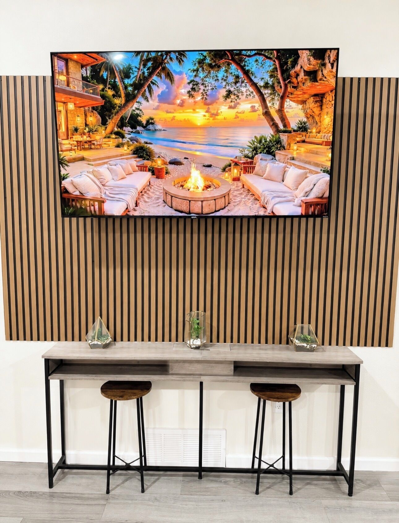A flat-screen TV mounted on a wall with a wooden slat design, a wooden bar table with two stools, and a fire pit in the center.