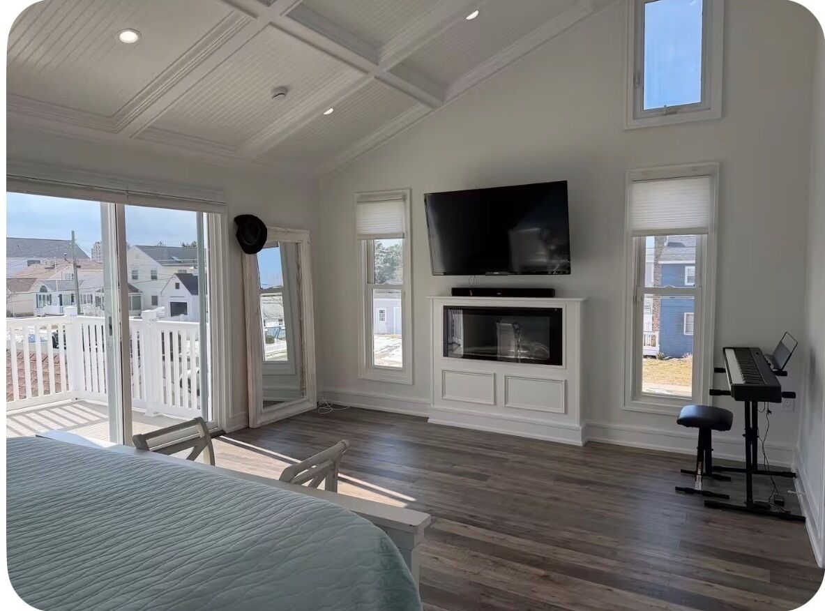A spacious room with a large bed, a television mounted above a fireplace, a piano, and a balcony with a view of houses.