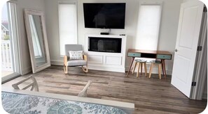 Ein Zimmer mit Kamin, einem darüber hängenden Fernseher, einem Schreibtisch mit zwei Stühlen und einem Fenster mit Außenblick.