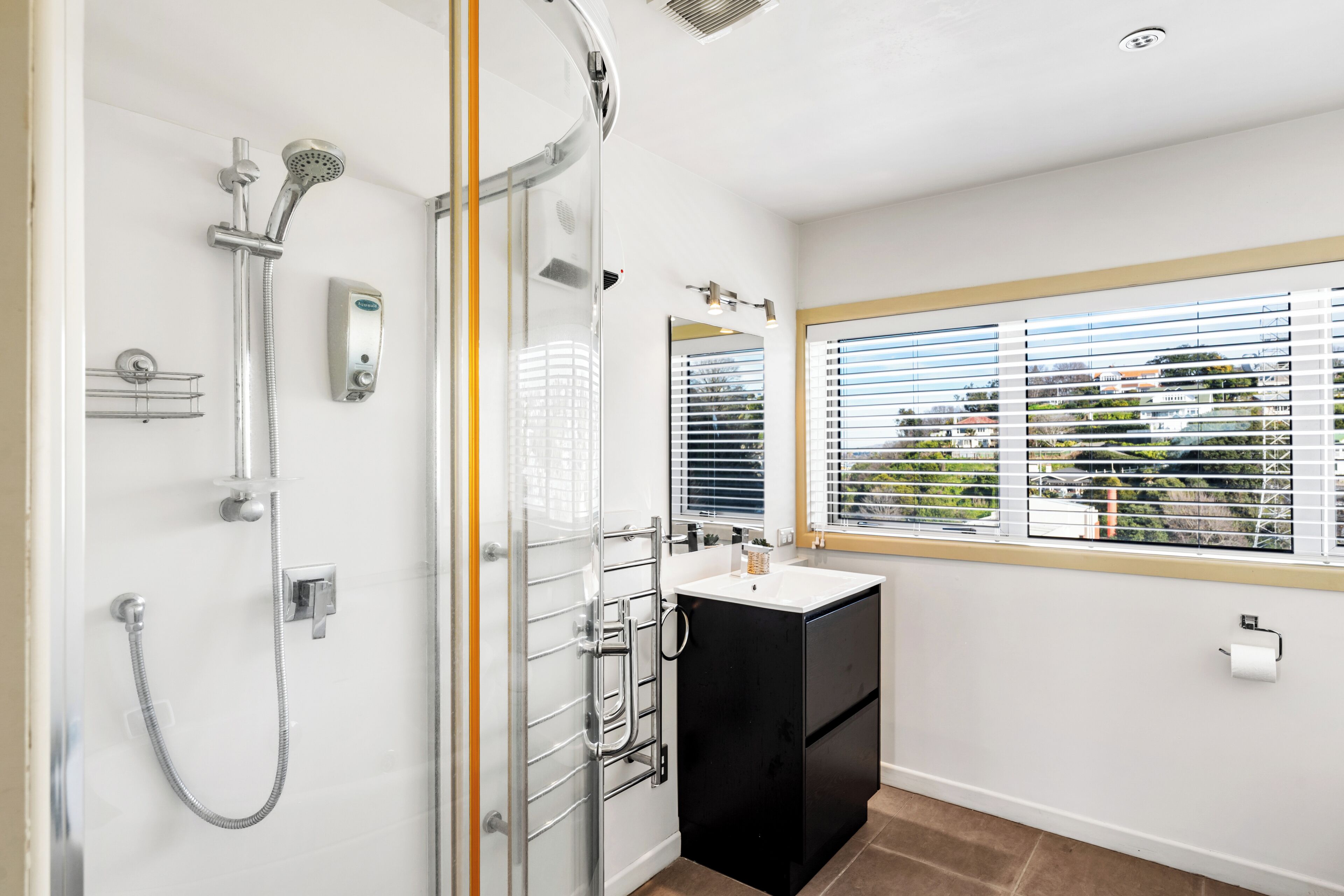 Ein Badezimmer mit Duschkabine aus Glas, einem weißen Waschbecken und einem Fenster mit Jalousien.