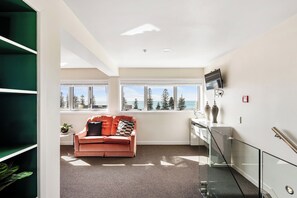 Una sala de estar moderna con un sofá, un televisor y ventanas amplias con vistas a árboles y al océano.