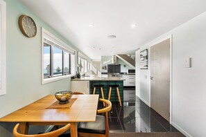 A modern kitchen with a central island, a dining area with a wooden table and chairs, and a view of the outdoors through large windows. - Sea and City Penthouse - Napier CBD (Napier)