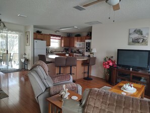 A modern living room with a sofa, a dining table with a decorative centerpiece, a kitchen area with wooden cabinets, a microwave, and a flat-screen TV.