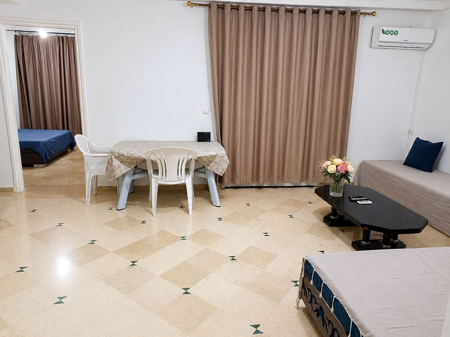 A room with a bed, a sofa, a table with a vase of flowers, a chair, a table with a remote control, and a curtain.