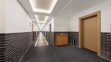 A long, narrow hallway with a wooden door on the right, a reception desk with a plant, and a tiled floor.