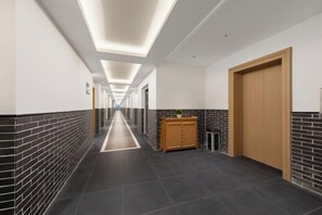 A long, narrow hallway with a wooden door at the end, a reception desk, and a trash bin.