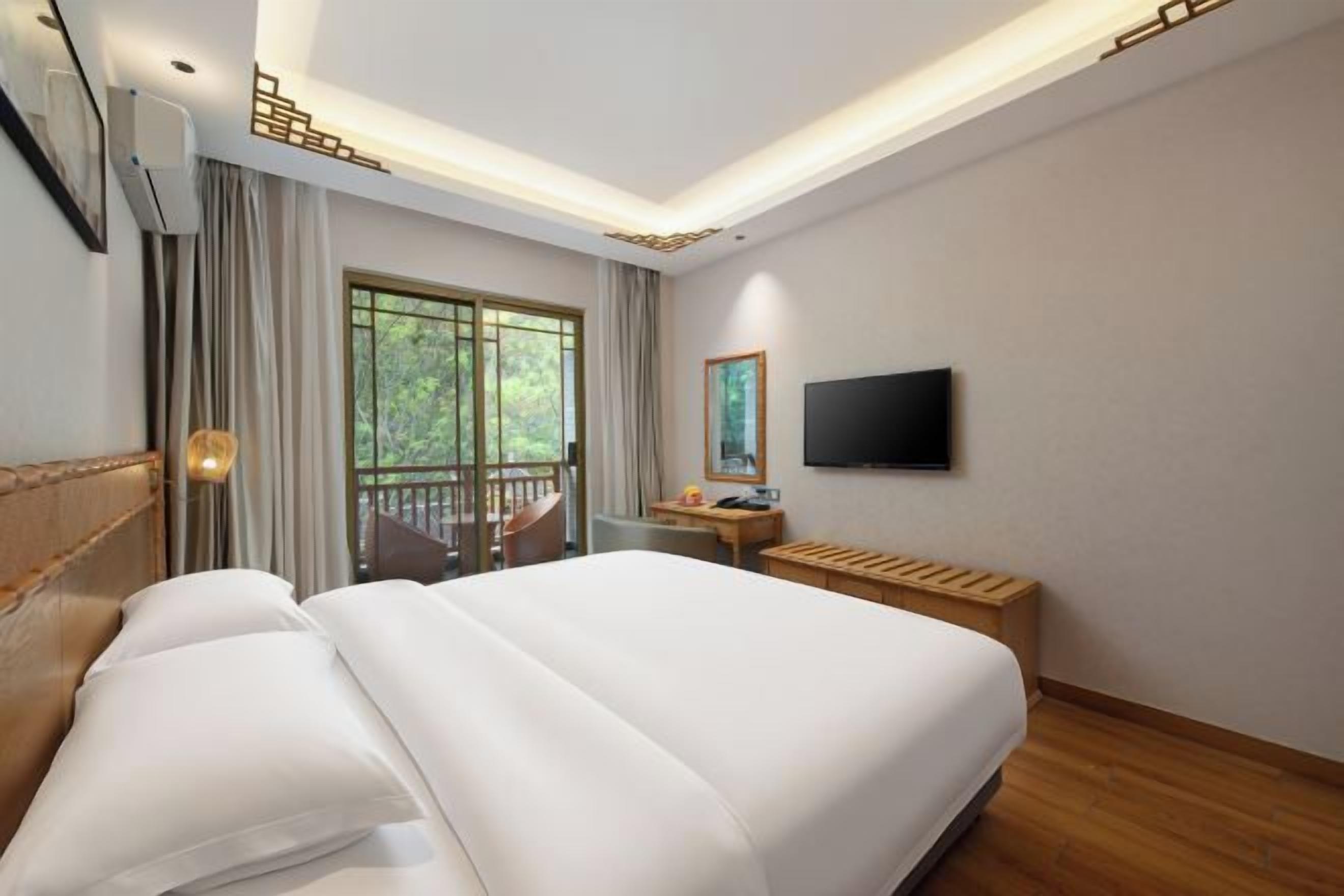 A hotel room with a large bed, a desk, a TV, and a balcony with a view of greenery.