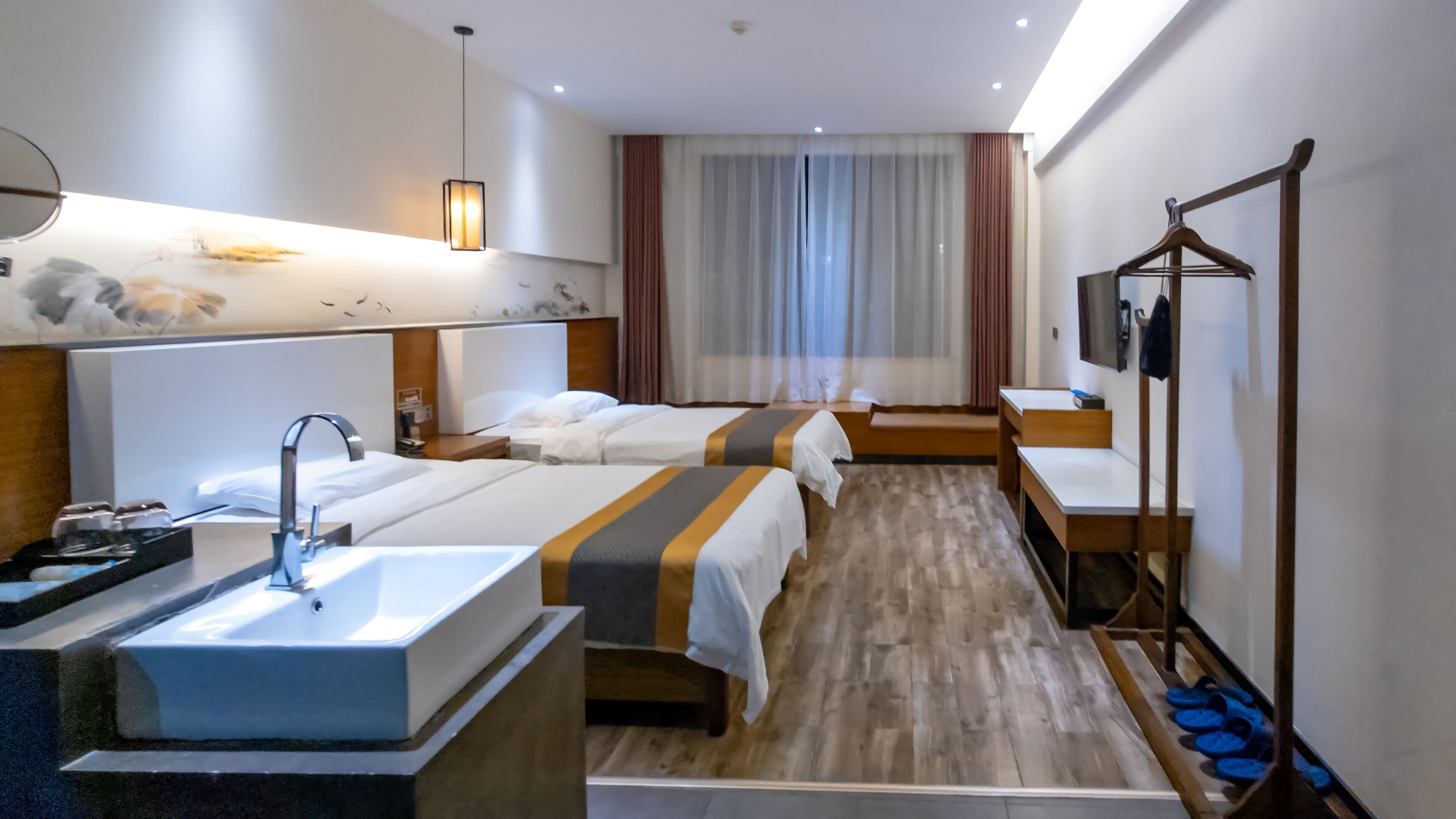 A hotel room with a large bed, a smaller bed, a desk, a vanity with a mirror, and a hanging light fixture.
