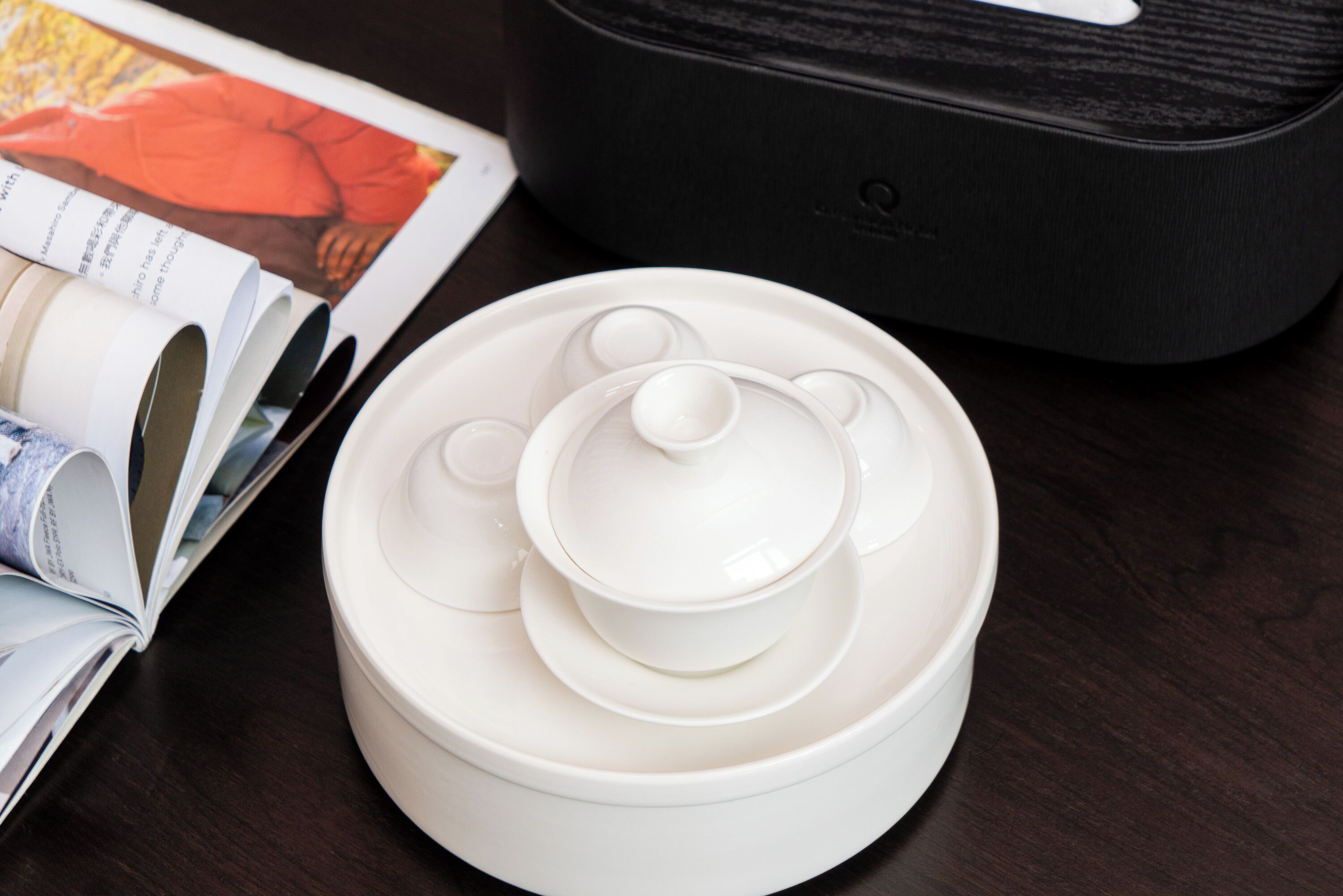 A stack of white ceramic containers with lids, a black appliance, and a magazine on a wooden surface.