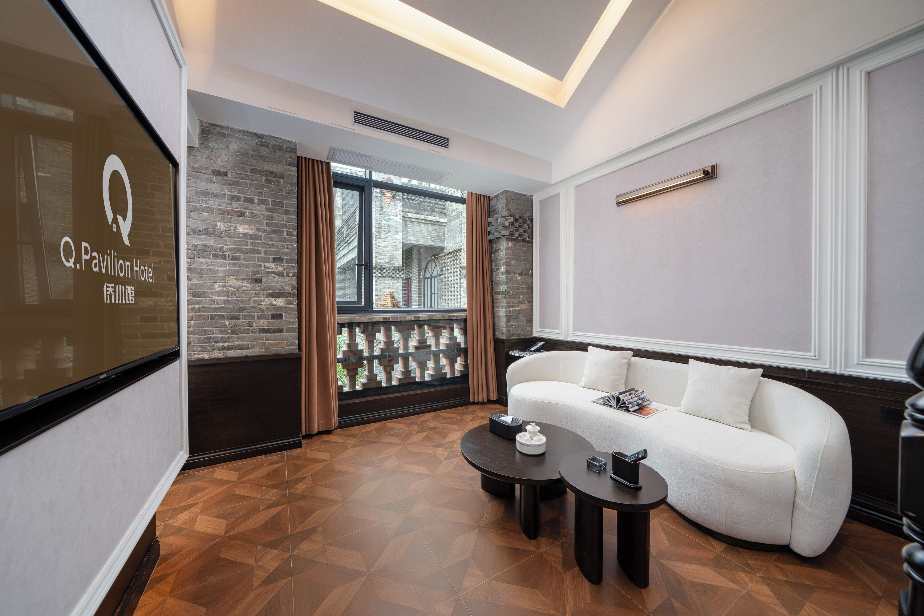 Habitación de hotel moderna con un sofá blanco, una mesa de centro y vista a un edificio de ladrillo a través de una puerta de vidrio.
