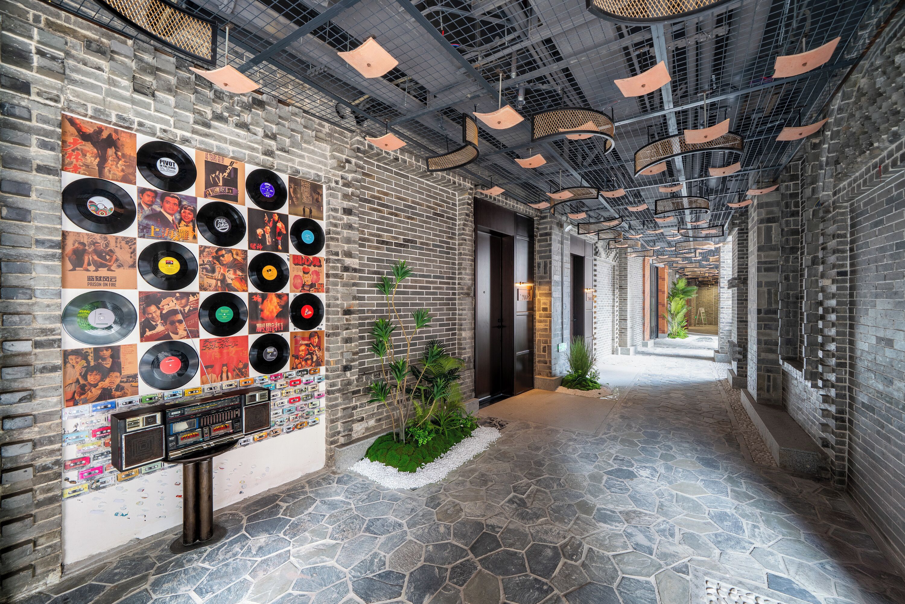 Un pasillo con un muro que exhibe discos de vinilo y cintas de cassette, suelo de baldosas de piedra y techo metálico con luces colgantes.