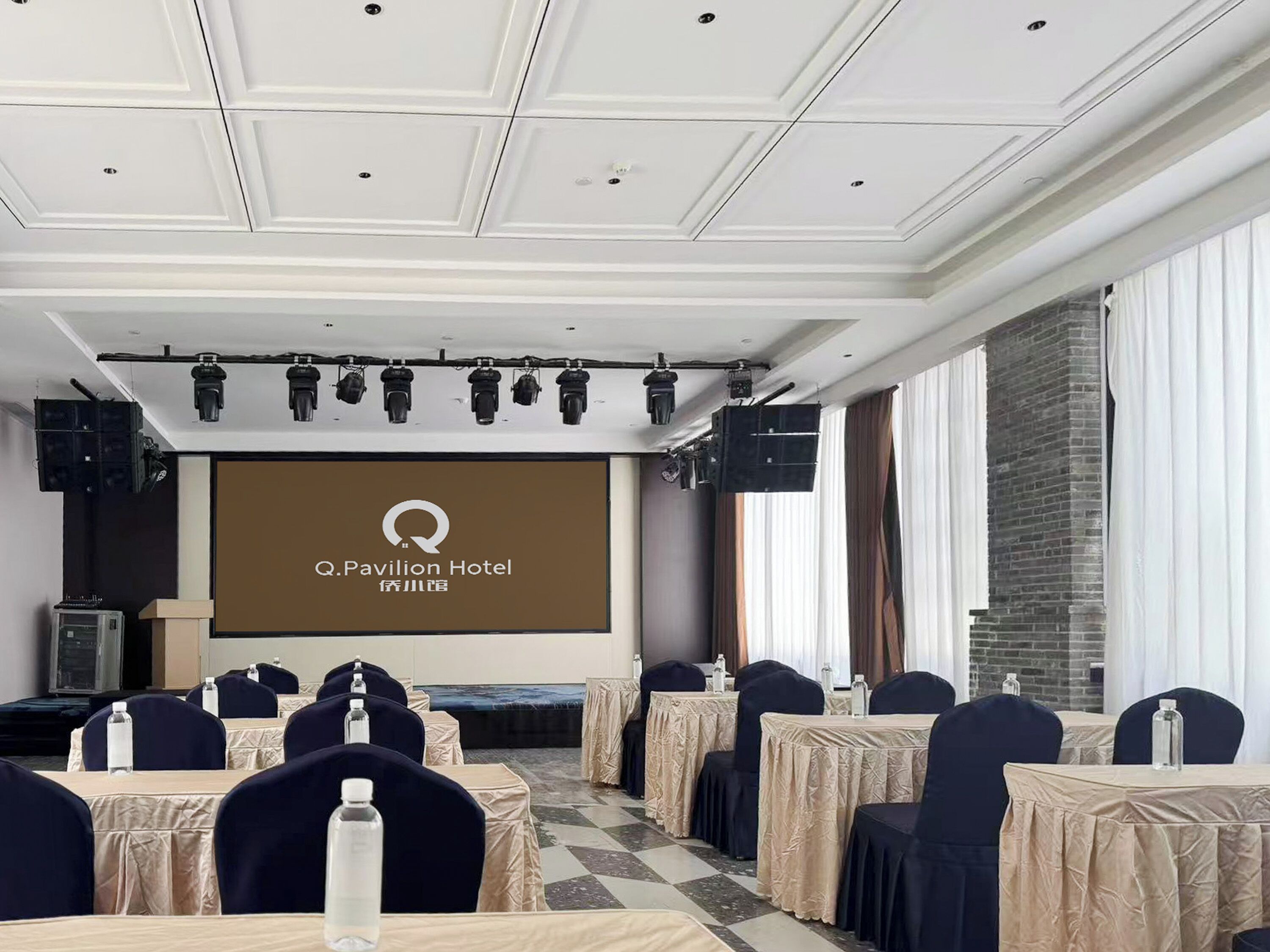 A conference room with a large screen displaying the Q Pavilion Hotel logo, multiple tables with chairs, and a brick wall.
