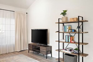 A living room with a flat-screen TV on a wooden stand, a bookshelf with various items, and a large window with curtains. - Tempe Coco-Ari 🌵⛳ Heated Pool, Firepit & Space for 9 in South Tempe (Tempe)