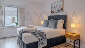Ein Schlafzimmer mit Bett, Nachttischlampen, einem Fenster mit Jalousien, einem Gemälde und einem kleinen Tisch mit Lampe.