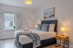 Ein Schlafzimmer mit Bett, Nachttischlampen, einem Fenster mit Jalousien, einem Gemälde und einem kleinen Tisch mit Lampe.