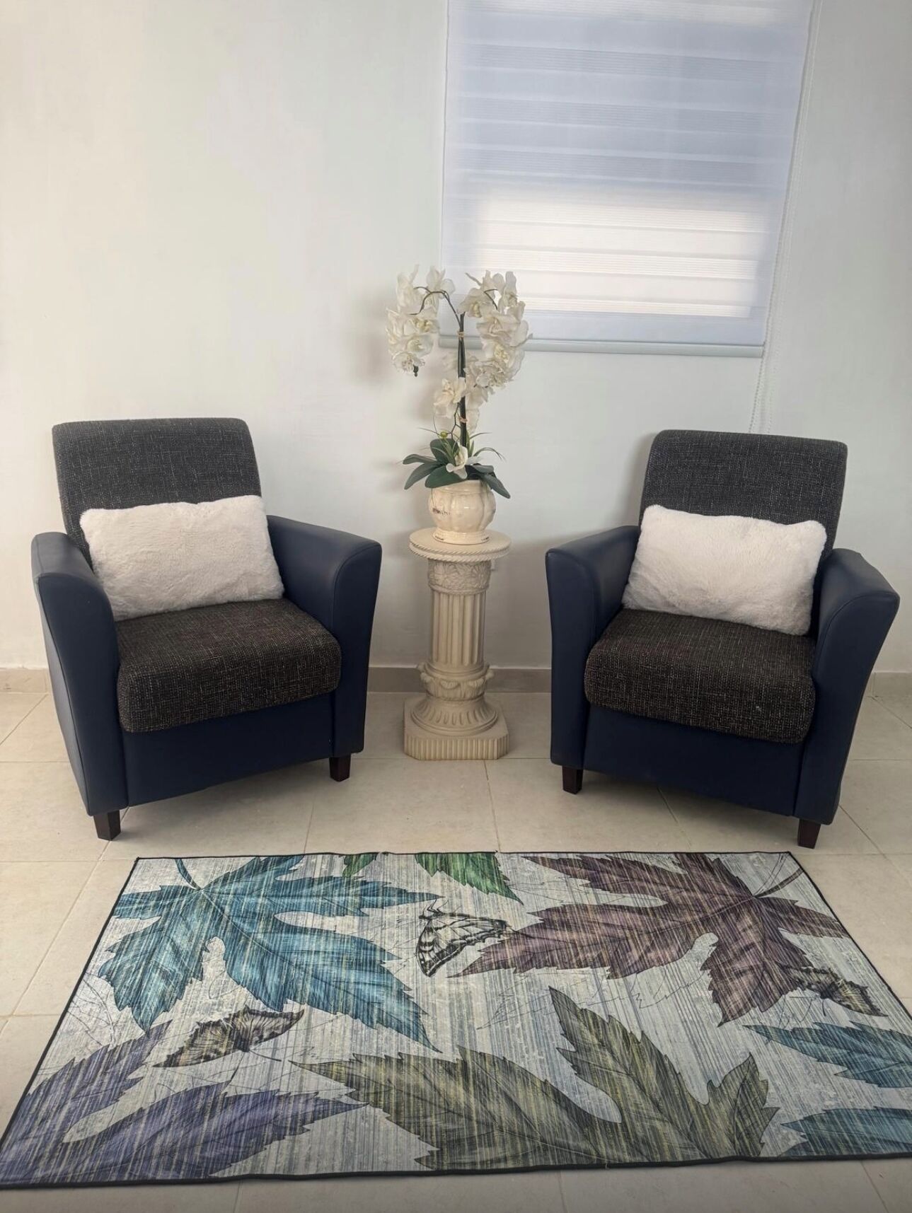 Two armchairs with white cushions, a small table with a potted orchid, and a patterned rug on a tiled floor.