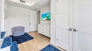 A room with a wooden floor, white walls, a white cabinet with drawers, a television on the wall, and a patterned ottoman. - NEW! Gulf View Suite | Gulf Views, Shared Pool, Exclusive Beach Access (Siesta Key)