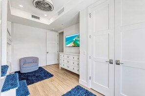 Ein Zimmer mit Holzboden, weißen Wänden, einem weißen Schrank mit Schubladen, einem an der Wand befestigten Fernseher und einem gemusterten Hocker.