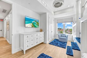 A modern hotel room with a large TV, white dresser, blue rugs, and a view of the outdoors. - NEW! Gulf View Suite | Gulf Views, Shared Pool, Exclusive Beach Access (Siesta Key)