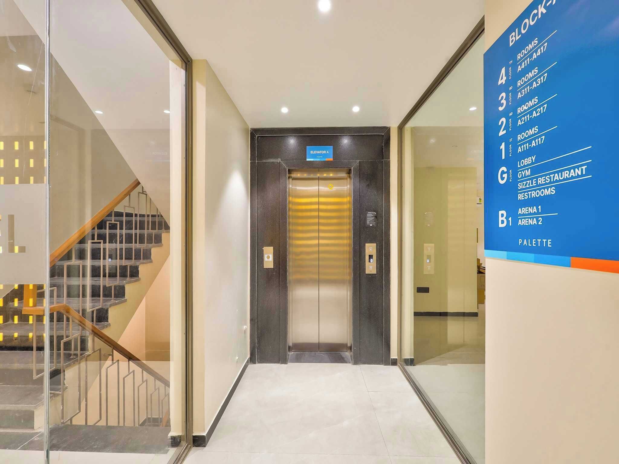 A hotel lobby with a staircase, an elevator, and a directory sign listing room numbers and amenities.