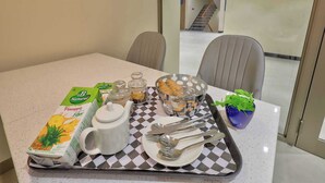 A breakfast setup with a pineapple juice box, a teapot, a bowl of pastries, and a small plant on a checkered tray. - Palette Convention Hotel Millennium City (Gurugram)