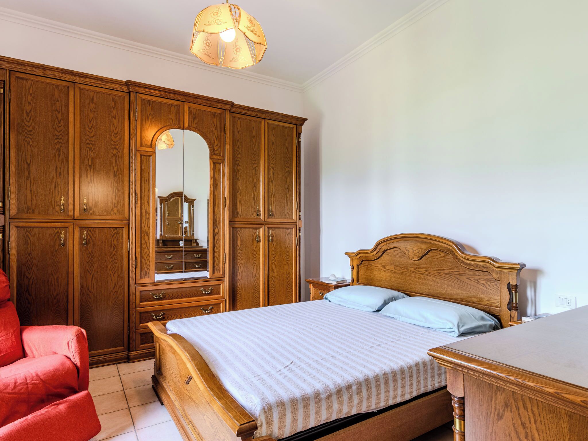 A bedroom with a wooden bed, a red chair, a wardrobe, and a mirror.