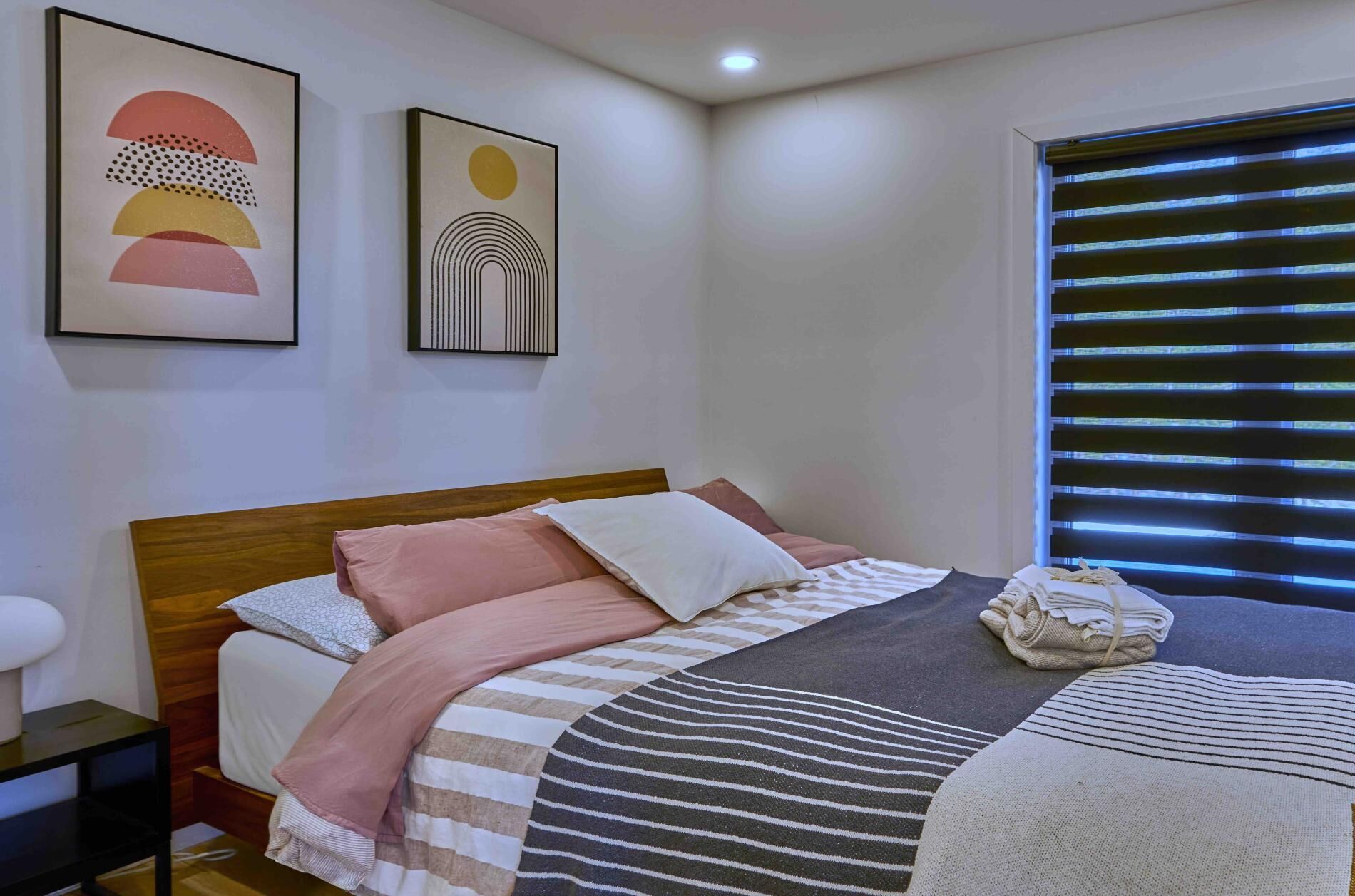 Un dormitorio con cama, almohadas, una colcha, una mesita de noche y dos cuadros en la pared.