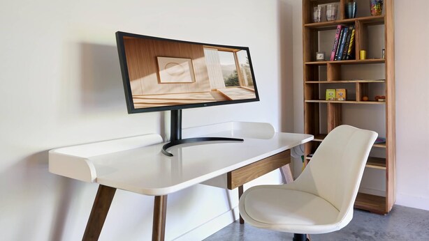Un escritorio moderno con un monitor de computadora, una silla blanca y una estantería de madera al fondo.