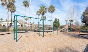 Ein Spielplatz mit Schaukeln, einer Kletterkonstruktion und einem eingezäunten Bereich mit Palmen und einem Gebäude im Hintergrund.