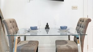 A modern dining area with a glass table, two cushioned chairs, a Buddha statue, and a television mounted on the wall. - Pet Friendly Studio in Fulham, Sleeps 2 (London)