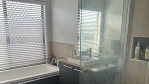 A modern bathroom with a glass shower enclosure, a window with blinds, a sink, and a small shelf with toiletries. - Room-our Little World in Caloundra (Sunshine Coast)