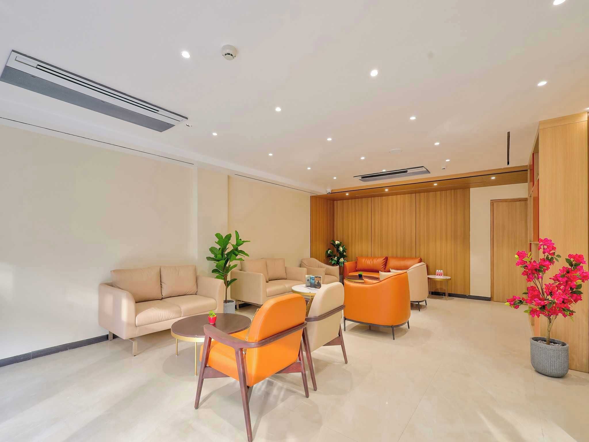 A modern hotel room with beige sofas, orange chairs, a small table, a potted plant, and a wooden panel wall.